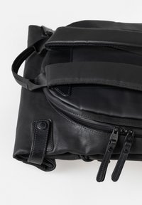 Zwarte opgevouwen rugzak met gevoerde banden, dubbele ritssluitingen en een klein handvat, geplaatst tegen een witte achtergrond.