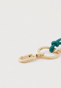 Kultainen metallilukko, jossa turkoosi punottu nyöri, pyöreä rengas ja logoteksti. Sileä pinta ja kiiltävä viimeistely.