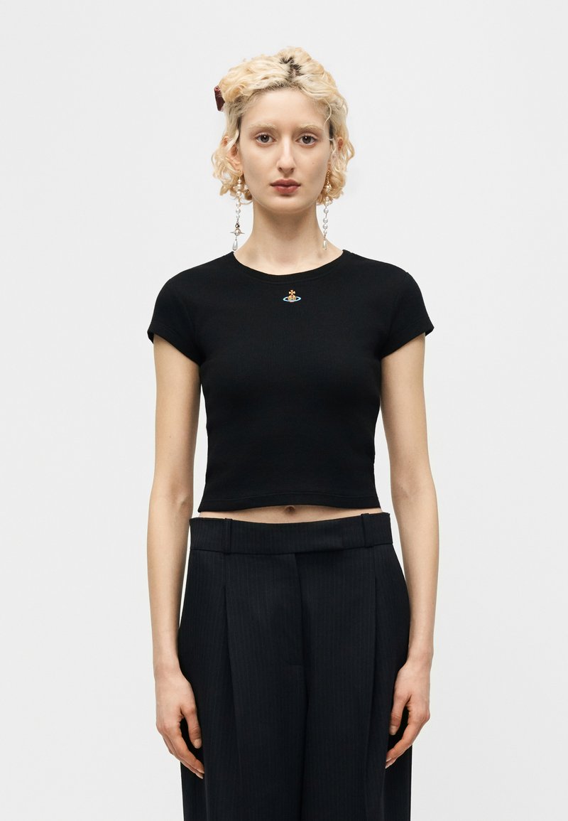 Νέος με κοντά σγουρά ξανθά μαλλιά φοράει μαύρο στενό κορμάκι με μικρό πολύχρωμο έμβλημα και μαύρο παντελόνι με ψηλή μέση.