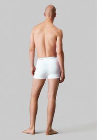 Boxers en coton blanc avec une ceinture élastique noire marquée, vus de dos. Le modèle est pieds nus sur un fond neutre.