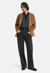 Vrouw in een bruine blazer over een zwart-grijs houndstooth hemd, zwarte high-waisted wijde jeans en zwarte schoenen, staand met haar handen in haar zakken.