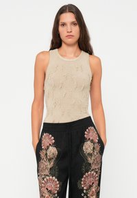 Woman with long brown hair wearing a sleeveless beige top with textured details and black pants with large pink floral embroidery.