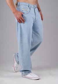 Man wearing light blue loose jeans and white sneakers, standing with one hand in pocket against plain background.