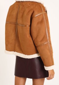 Brązowa zamszowa kurtka z kremowym futrem syntetycznym i skórzanymi akcentami; posiada pudełkowy kształt i widoczne detale szwów.