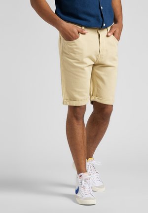 Shorts beiges en denim jusqu'aux genoux, avec ourlets retroussés et poches avant. Associés à des baskets montantes blanches avec des accents bleus.