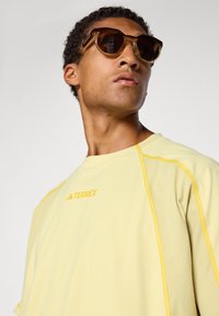 Yellow athletic t-shirt with short sleeves, featuring contrasting yellow stitching and the "TERREX" logo. Complemented by brown sunglasses.