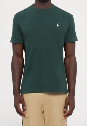 T-shirt vert foncé à manches courtes avec un petit logo blanc sur la poitrine gauche, porté avec un pantalon cargo beige.