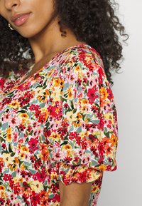 Blusa floral com mangas curtas bufantes, apresentando uma estampa multicolorida de vermelho, rosa, laranja e verde sobre um fundo claro.