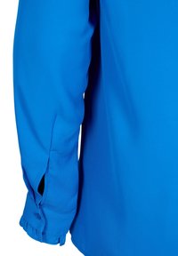 Blouse bleue à manches longues, fabriquée en tissu lisse. Elle présente un poignet boutonné et une fente sur le côté, mettant en valeur des lignes épurées et une couleur vive.