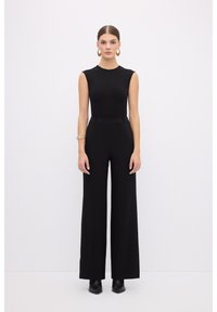 Bodysuit sans manches noir associé à un pantalon large taille haute. La mannequin porte des bottines noires et des boucles d'oreilles en or. Texture lisse.