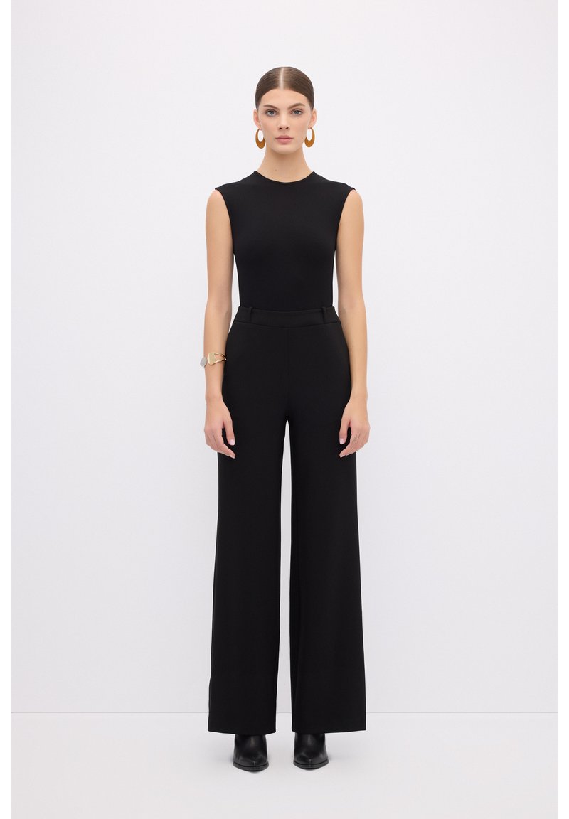 Bodysuit sans manches noir associé à un pantalon large taille haute. La mannequin porte des bottines noires et des boucles d'oreilles en or. Texture lisse.
