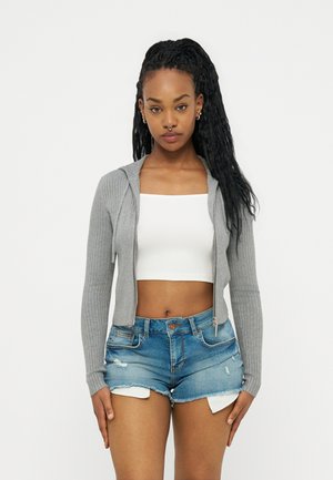 Femme aux longs cheveux tressés portant un sweat à capuche gris zippé, un crop top blanc et un short en denim déchiré, debout devant un fond uni.
