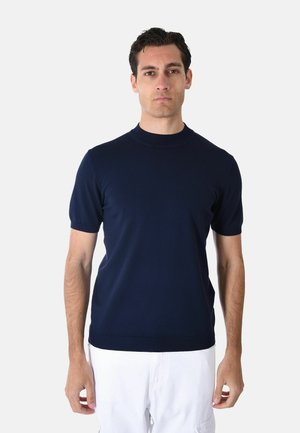 Uomo con indosso una maglietta blu navy a maniche corte con collo tondo e pantaloni bianchi, in piedi davanti a uno sfondo grigio chiaro uniforme.