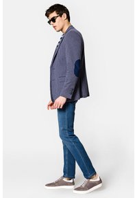 Granatowa marynarka w kratkę z kontrastującymi granatowymi łatami na łokciach, w połączeniu z niebieskimi dżinsami i szarymi trampkami. Dodatki obejmują czarne okulary przeciwsłoneczne.