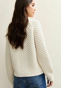 Kremowy sweter z dzianiny o fakturowanym wzorze, okrągłym dekolcie i ribbowym wykończeniu, w połączeniu z niebieskimi dżinsami.