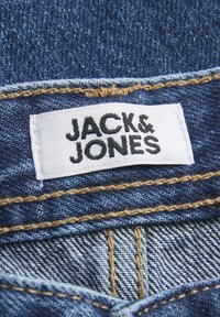 Étiquette en tissu denim avec "JACK & JONES" imprimé en texte noir gras sur fond blanc, cousue sur du denim bleu brut avec des accents de fil doré.
