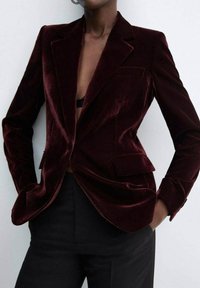 Veste en velours bordeaux avec une coupe ajustée, un design à simple boutonnage, des revers crantés et des poches avant. Texture lisse et forme structurée.