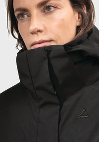 Schwarze Jacke mit hohem Kragen, aus glattem Stoff und mit einem gestickten Logo auf der linken Seite. Insgesamt elegantes und minimalistisch gestaltetes Design.