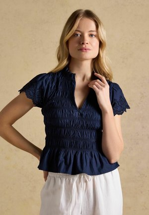 Blonde Frau trägt eine marineblaue Bluse mit kurzen Ärmeln, gerafften Details und Ärmeln aus Lochspitze, kombiniert mit weißen Hosen.