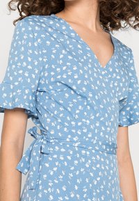 Blå blommig omlottklänning med korta volangärmar, med V-ringning, lätt tyg och vita blommönster över hela plagget.