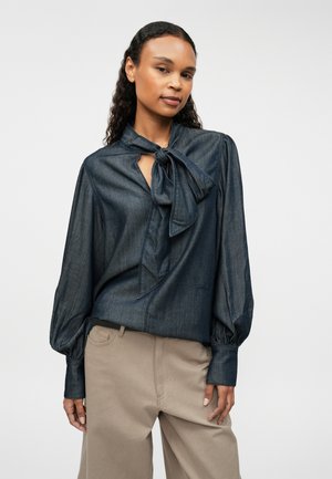 Dunkelblaue Denim-Bluse mit einem gebundenen Kragen, langen Puffärmeln und einer Schlüssellochöffnung am Ausschnitt. Kombiniert mit hellbeigen Hosen.