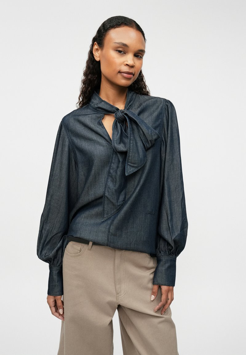 Tumši džinsi blūze ar sasietu apkakli, garām pūkainām piedurknēm un atslēgvārtiņu atvēršanu pie kakla. Saskaņota ar gaiši bežām biksēm.