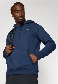 Marineblå hettegenser laget av mykt stoff, med en frontlomme (kangaroo pocket), snøring i hetten og en liten logo på brystet.