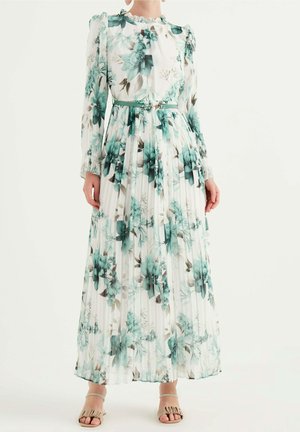 Robe blanche plissée à manches longues avec imprimé floral turquoise, col à volants, ceinture assortie, portée avec des sandales ouvertes beiges.