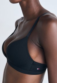 Black, triangle-shaped bra made of smooth fabric, featuring thin straps and a small logo patch with red, white, and blue accents.