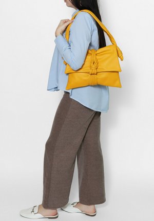 Gelbe Lederhandtasche mit einem geknoteten Akzent, flacher Form und dickem Schultergurt. Das Model trägt eine hellblaue Bluse und braune weite Hosen.