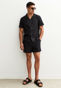 Black short-sleeve button-up shirt with textured pattern and single chest pocket, paired with matching shorts and black sandals.