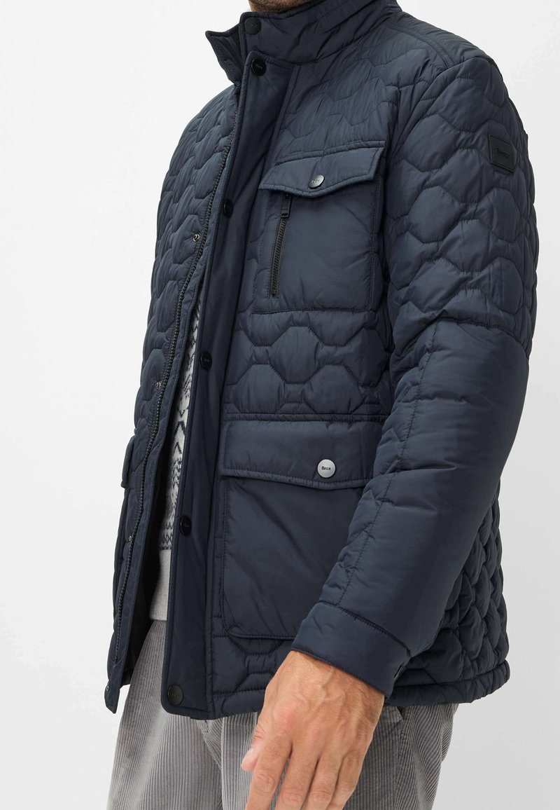 Homme portant une veste matelassée bleu foncé avec plusieurs poches et boutons, debout devant un fond clair uni.