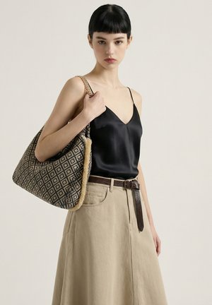 Jeune femme aux cheveux courts et foncés porte un caraco en satin noir, une jupe beige taille haute avec une ceinture marron, portant un grand sac à épaule à motifs.