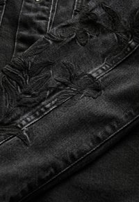 Țesătură de denim negru cu broderie florală neagră complexă pe cusătura laterală, cu o textură netedă și detalii vizibile de cusătură.