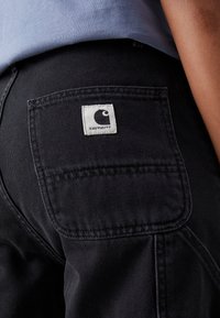 Zwarte denim broek met een achterzak die een rechthoekig wit label met een zwart logo bevat. De stof heeft een stevige, gestructureerde uitstraling.