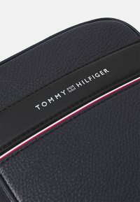 Ciemnoniebieska skórzana saszetka teksturowana z czarnymi akcentami i czerwoną oraz białą paskiem. Zawiera logo "TOMMY HILFIGER" w srebrnym napisie.