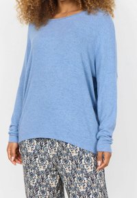 Frau trägt einen lockeren, hellblauen Pullover mit langen Ärmeln und gemusterte Hosen mit blau-beigem Blumendesign.