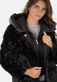 Cappotto in pelliccia sintetica nera con un morbido strato interno trapuntato nero, grande cappuccio, zip dorata e una texture distintiva e soffice.