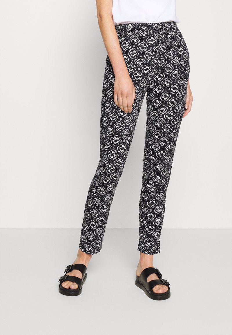 Personne portant un pantalon slim noir à motifs, un haut blanc et des sandales noires à double bride, debout devant un fond uni.
