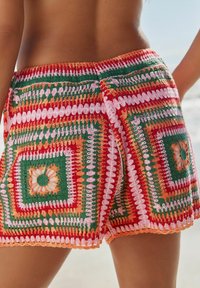 Hæklede shorts med et farverigt geometrisk mønster i rød, grøn, orange og pink, med dekorative accenter og bølgede kanter.