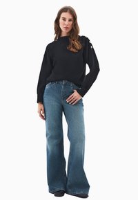 Schwarzer Strickpullover mit Bootsausschnitt, mit Knopfdetails an den Ärmeln, kombiniert mit weit geschnittenen blauen Jeans aus Denim mit einem ausgewaschenen Finish.