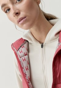 Rosa pufferväst med en mjuk grå insida som har upprepande röda grafiska mönster. Dragkedja framtill och en fastsydd huvtröja under.