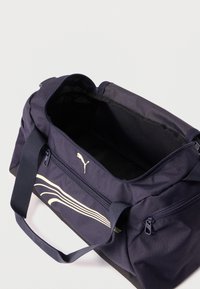 Σκούρο μπλε αθλητική τσάντα Puma με το κύριο φερμουάρ ανοιχτό, μπροστινή τσέπη με φερμουάρ και λευκό σχέδιο κυμάτων, και λαβές μεταφοράς.