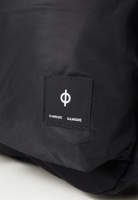 Gros plan sur un tissu noir avec un patch carré cousu arborant un symbole circulaire blanc et le texte "SAMSØE SAMSØE."