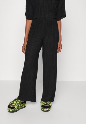 Unterkörper einer Person, die lockere schwarze plissierte Hose und neongrüne und schwarz zebragestreifte Plateau-Sandalen mit schwarzen Socken trägt.
