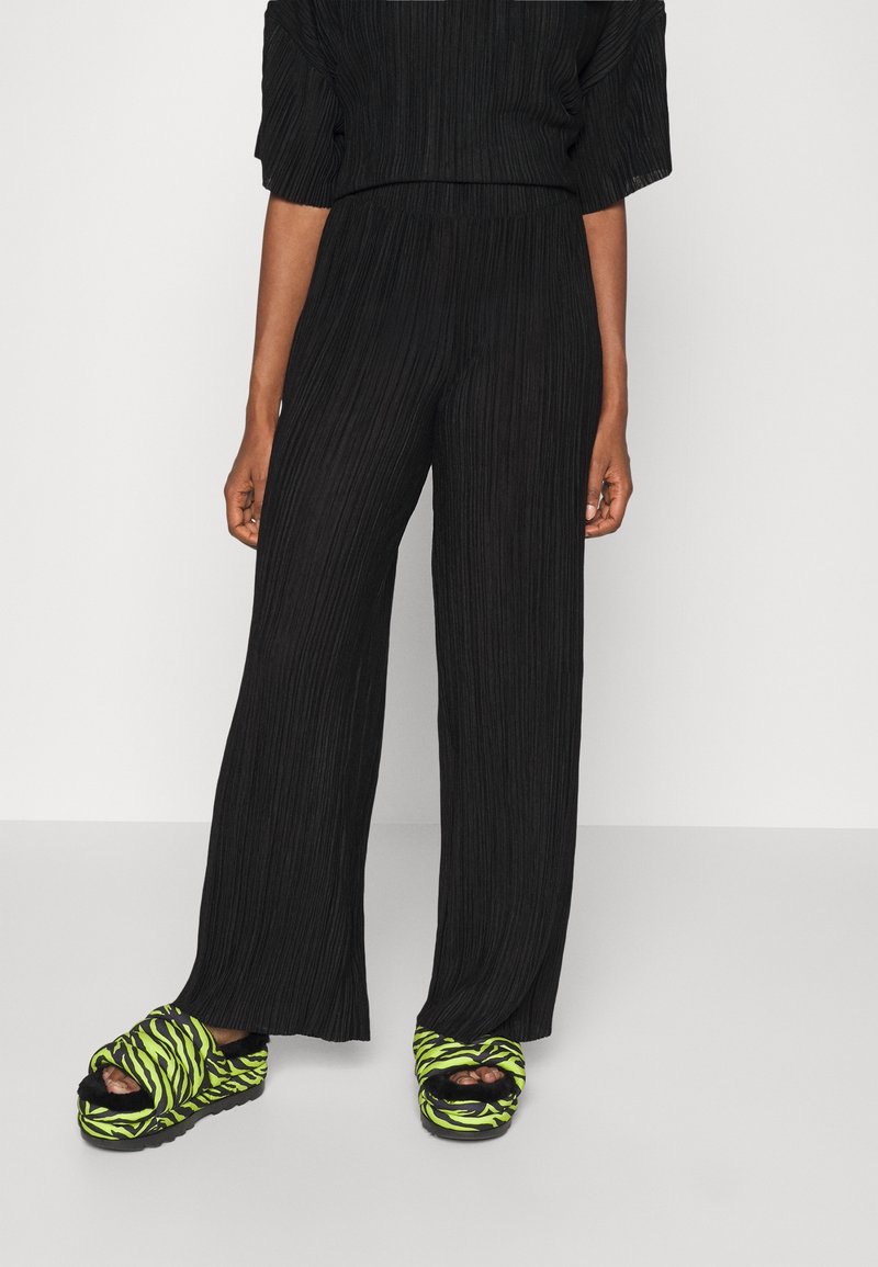 Partie inférieure du corps d'une personne portant un pantalon noir plissé ample et des sandales compensées à rayures zébrées vert fluo et noir avec des chaussettes noires.