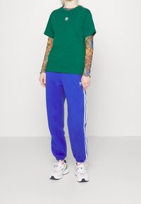 Vihreä puuvillapaita, jossa on valkoinen logo, kerroksittain monivärisen kuvioidun pitkähihasen päällä. Siniset verkkarit, joissa on valkoiset sivusuorat ja logo.