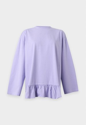 Femme portant un haut lavande oversize avec un ourlet à volants, un jean bleu et des baskets bleu marine, debout de profil sur fond blanc.