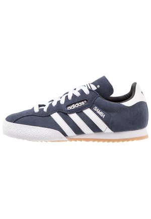 Granatowy sneaker Adidas Samba z białymi paskami, białymi sznurowadłami, zamszową cholewką i gumową podeszwą, skierowany w lewo.
