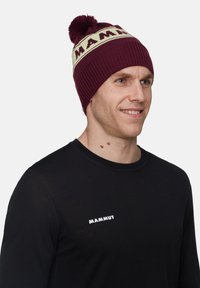 Vinröd stickad mössa med en beige bård som har "MAMMUT" i fet stil och en pom-pom på toppen. Ribbade detaljer.